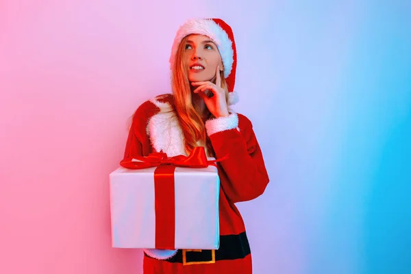 Pensive dreaming girl in Santa hat with Christmas gift standing on background with neon light — Stock Photo, Image