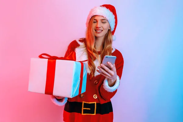 Happy young woman in Santa hat, with Christmas gifts, uses smartphone — Stock Photo, Image