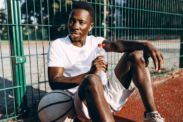 Afro-américain se repose après une séance d'entraînement avec une bouteille d'eau et écoute de la musique dans les écouteurs — Photo