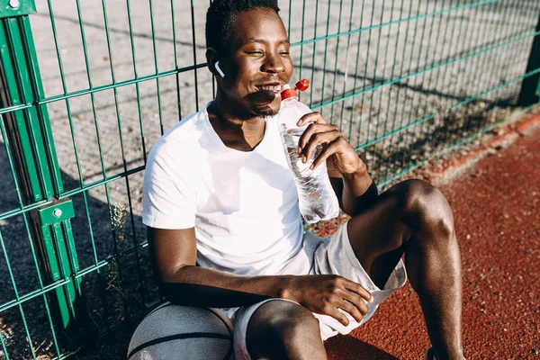 Afro-américain se repose après une séance d'entraînement avec une bouteille d'eau et écoute de la musique dans les écouteurs — Photo