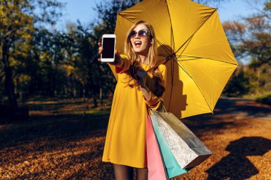 Alışveriş torbaları taşıyan mutlu bir kız, boş bir cep telefonu ekranı gösteriyor.