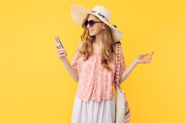 Yazlık şapkalı ve güneş gözlüklü mutlu bir genç kadın izole edilmiş sarı bir arka planda akıllı bir telefon tutuyor.