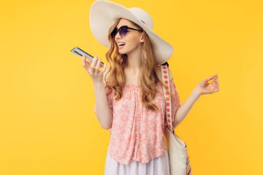 Yazlık şapkalı ve güneş gözlüklü mutlu bir genç kadın izole edilmiş sarı bir arka planda cep telefonuyla konuşuyor.