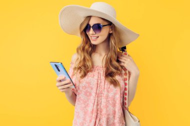 Yazlık şapkalı ve güneş gözlüklü mutlu bir genç kadın izole edilmiş sarı bir arka planda akıllı bir telefon tutuyor.