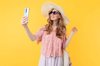 Yazlık şapkalı ve güneş gözlüklü mutlu bir genç kadın izole edilmiş pembe bir arka planda cep telefonuyla selfie çekiyor.