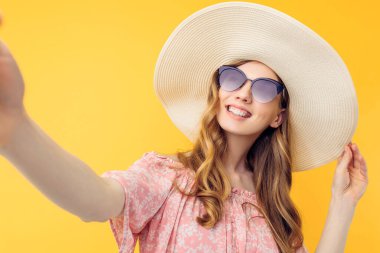 Yazlık şapkalı ve güneş gözlüklü mutlu bir genç kadın izole edilmiş sarı bir arka planda cep telefonuyla selfie çekiyor.