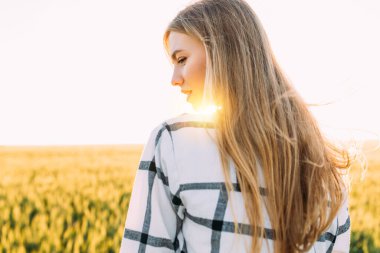 Arkaya bakan, işaretli tişörtlü mutlu bir kadın güneşli bir günbatımında buğday tarlasında geziniyor.