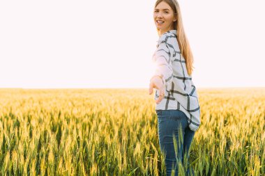 Arkaya bakan, işaretli tişörtlü mutlu bir kadın güneşli bir günbatımında buğday tarlasında geziniyor.