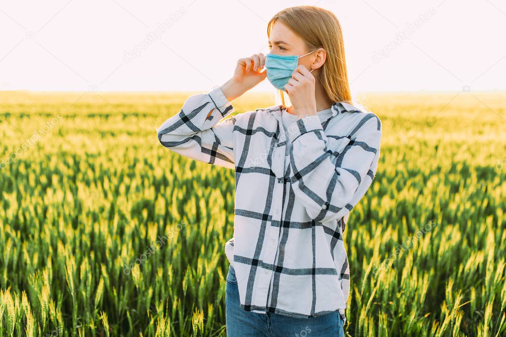 beautiful woman in a checked shirt and a protective medical mask on her face stands in a wheat field at sunset. Quarantine, coronavirus, crop
