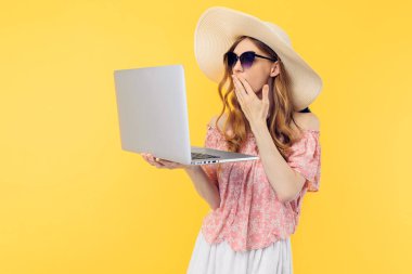 Hasır şapkalı ve güneş gözlüklü güzel bir kadının sarı arka planda dizüstü bilgisayar kullanmasına şaşırdım.