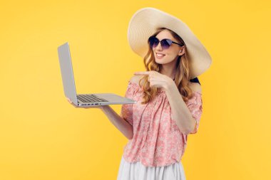 Sarı arka planda dizüstü bilgisayar kullanan hasır şapkalı ve güneş gözlüklü çekici genç bir kadın.
