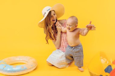 Yazlık şapkalı mutlu genç bir anne küçük bir çocuk ve sarı bir arka planda şişme bir yüzme çemberiyle oturur.