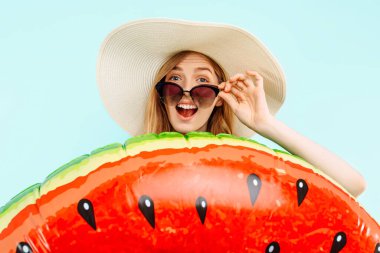 Şok olmuş ve şaşırmış güneş gözlüklü bir kız ve elinde mavi arka planda şişme bir yüzük tutan bir yaz şapkası.