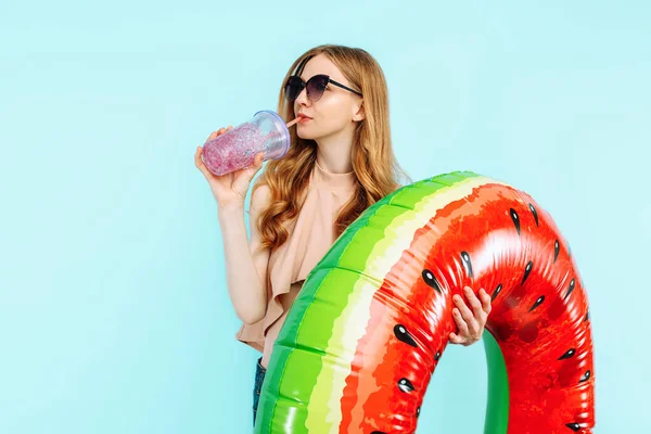 身穿泳衣 戴着太阳镜的快乐漂亮的女孩拿着鸡尾酒和一个蓝色背景的充气游泳圈 — 图库照片