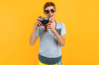 Happy smiling teen guy in sunglasses stands with a camera on a yellow background clipart