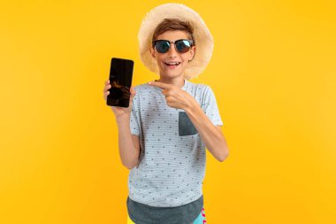 Yazlık şapkalı mutlu genç adam, boşluğun bir kopyası için boş bir cep telefonu ekranı gösteriyor, izole edilmiş sarı arka planda.