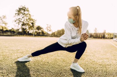 Yüzünde tıbbi koruyucu maske takan atletik genç bir kadın, dışarıda egzersiz yapıyor.