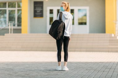 Tıbbi koruyucu maskeli genç bir kız, elinde bir çantayla bir binanın arka planında, açık havada duruyor. Karantina, koronavirüs