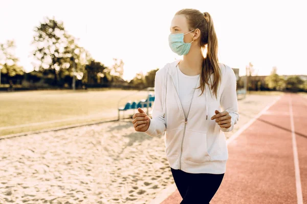 スポーツウェアを着たスポーティな若い女性と彼女の顔の医療保護マスクは 夜明けにスタジアムの周りを走っています スポーツ コロナウイルス — ストック写真