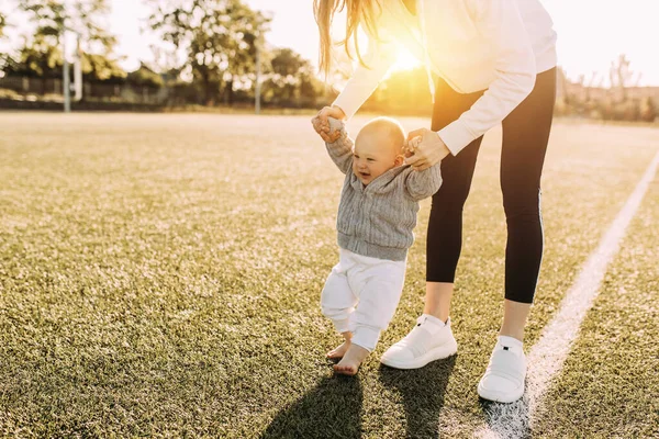 A mother teaches her young son to walk, holding his hands, in the fresh air against the background of dawn