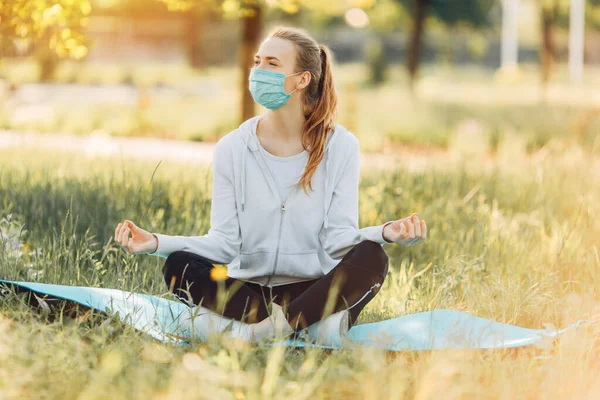 医療保護マスクの女の子は夜明けに緑の草の上のロータスの位置で瞑想します 健康的なライフスタイルとリラクゼーション — ストック写真