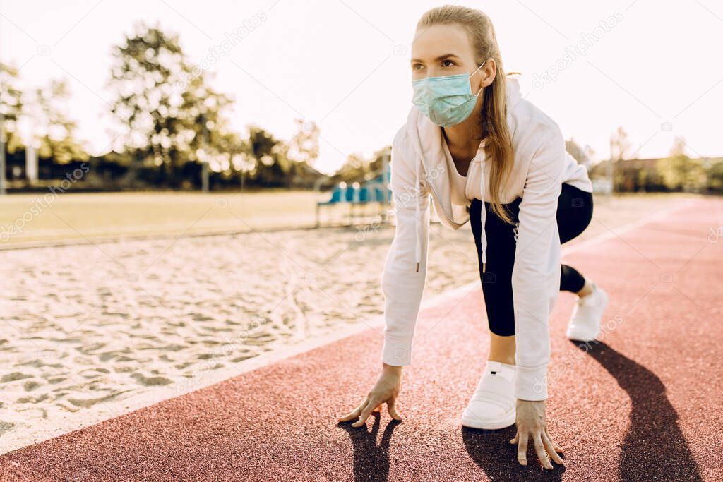 Athletic woman in sports clothing and a medical protective mask on her face, at the start line in the race. A female runner started the sprint from the starting line against the background of dawn.