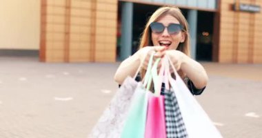 Güneş gözlüklü mutlu genç bir kadın, alışveriş torbalarıyla mutlu, şehirdeki bir alışveriş merkezinin arka planında duruyor.