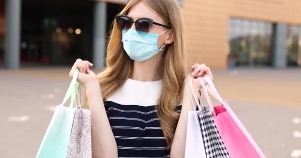 Jonge vrouw met een medisch beschermend masker op haar gezicht en zonnebril, met boodschappentassen, op straat op de achtergrond van een winkelcentrum — Stockvideo