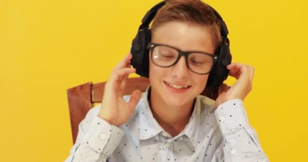 Feliz colegial emocionado escuchando música en los auriculares, sobre un fondo amarillo aislado — Vídeos de Stock