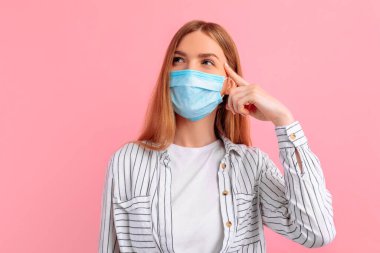 Yüzünde tıbbi koruyucu maske takan, soyutlanmış pembe arka planda düşünceli genç bir kadın. Koronavirüs salgını, karantina, virüs koruması