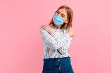 Tıbbi koruyucu maskeli genç ve çekici bir kadın soyutlanmış pembe bir arka plana karşı kendini kucaklıyor. Kendini sev.