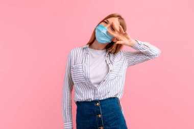 Yüzüne koruyucu maske takmış neşeli bir genç kadın resmi. Günlük kıyafetler giymiş, gülümsüyor ve stüdyoda pembe bir arka planda iki parmağı izole edilmiş bir barış işareti gösteriyor.