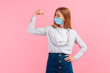 Yüzünde tıbbi koruyucu bir maske takmış, kollarında pembe bir arka planda pazıları olan mutlu ve güçlü genç bir kadın.