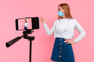 Yüzünde tıbbi koruyucu bir maske takan genç bir bayan öğretmen, uzaklardan öğrenme ya da cep telefonuyla pembe bir arka planda çevrimiçi dersler alıyor.