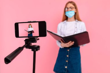 Yüzünde tıbbi koruma maskesi olan genç bir iş kadını çevrimiçi eğitim veriyor ya da cep telefonuyla pembe bir arka planda iş kursuna gidiyor.