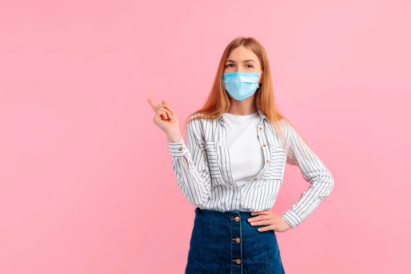 Yüzünde tıbbi koruyucu maske takan çekici genç bir kadın, parmağını telif boşluğuna gösteriyor, izole edilmiş pembe bir arka planda.