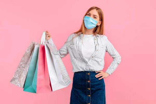 happy girl in a medical mask on her face, posing with shopping bags and looking at the camera on a pink background. Shopping, quarantine