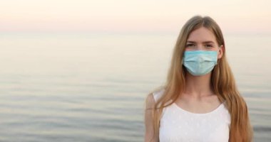  Beyaz elbiseli ve yüzünde tıbbi koruyucu bir maske olan güzel genç bir kadın kumsalda dinleniyor ve mavi denize bakan kameraya bakıyor.