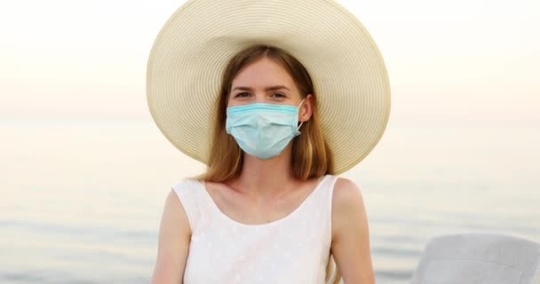 Bella giovane donna in un cappello estivo e maschera protettiva medica sul suo viso sulla spiaggia, godendo di una vacanza. Il concetto di vacanza estiva, quarantena, coronavirus — Video Stock