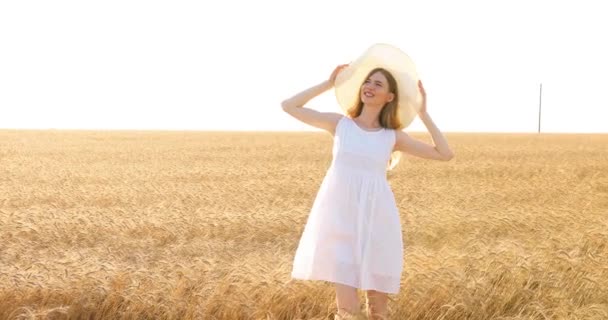 Mujer joven con un vestido blanco y sombrero de paja caminando por un campo de trigo al atardecer — Vídeos de Stock