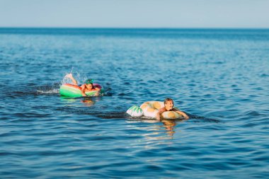 Çocuklar erkek ve kız, şişme dairelerle denizde yüzüyor, yaz tatillerinde kumsalda eğleniyorlar.