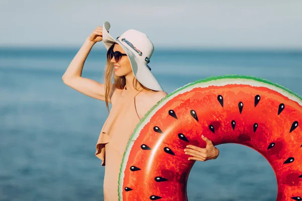 一个戴着夏帽 穿着泳衣 躺在海滩上 望着远方 手里拿着一个充气的游泳圈的漂亮姑娘的画像 — 图库照片