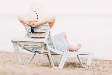 Yaz şapkalı, kumlu bir plajda dinlenen kız şezlongda uzanıyor. Yaz tatili, seyahat.
