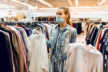 Yüzüne koruyucu bir maske takmış genç bir kadın kıyafet seçiyor ve bir giyim mağazasından alışveriş yapıyor. Alışveriş, alışveriş merkezi.