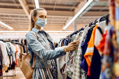 Yüzünde tıbbi koruyucu maske olan pozitif bir kız mağazadan yeni kıyafetler seçiyor ve gülümsüyor. Alışveriş, karantina, koronavirüs.
