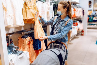 Bebek arabalı güzel bir kadın, yüzünde tıbbi koruyucu maske olan genç bir anne, bir çocuk mağazasından çocuk kıyafetleri seçiyor. Karantina, koronavirüs, alışveriş.
