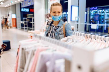 Yüzüne koruyucu bir maske takmış genç bir kadın kıyafet seçiyor ve bir giyim mağazasından alışveriş yapıyor. Alışveriş, alışveriş merkezi.