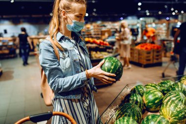 Yüzünde tıbbi koruyucu maskeli bir kadın süpermarketten meyve alıyor. Genç bir bayan markette yeşil bir karpuz tutuyor. Satış, alışveriş, yemek, koronavirüs.