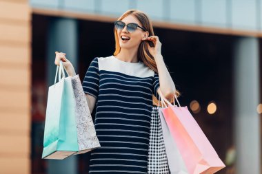 Güneş gözlüklü ve çok renkli alışveriş çantalı genç bir kadın alışveriş merkezinin önünde caddeden alışveriş yapıyor. Alışveriş, moda.