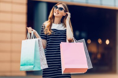 Güneş gözlüklü şık bir kız, alışveriş torbaları elinde, şehirde bir alışveriş merkezinin arka planında.